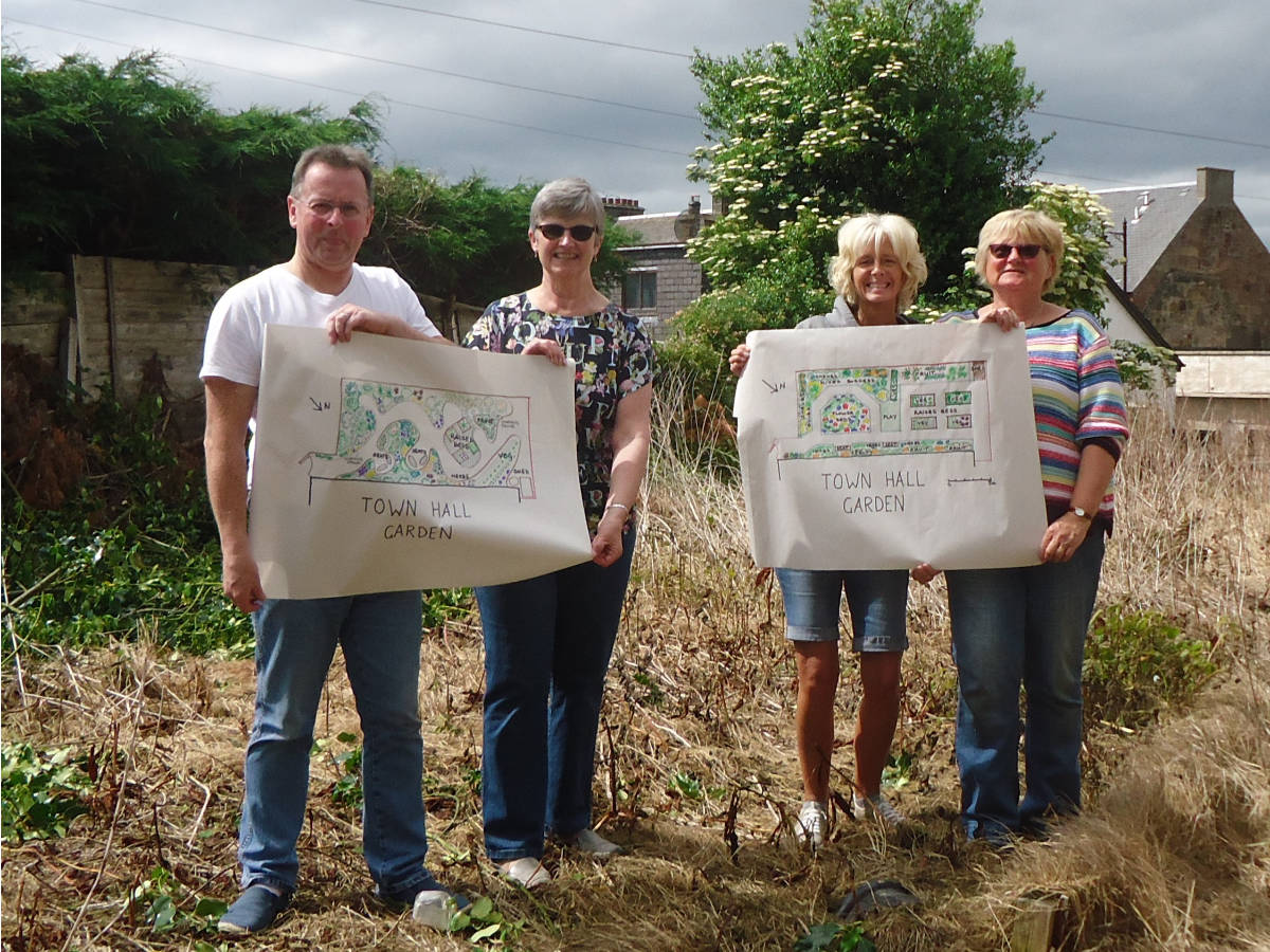 A photo of the garden site prior to work starting. Photo includes four people holder printed plans for the garden development.